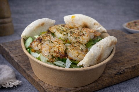 Spiced shrimp banh mi sandwich in a cardboard container with cucumber and fresh herbs on a wooden board