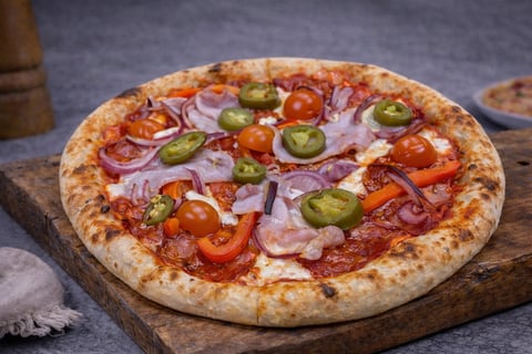 A freshly baked pizza on a wooden board topped with red peppers, jalapeños, ham, and cheese