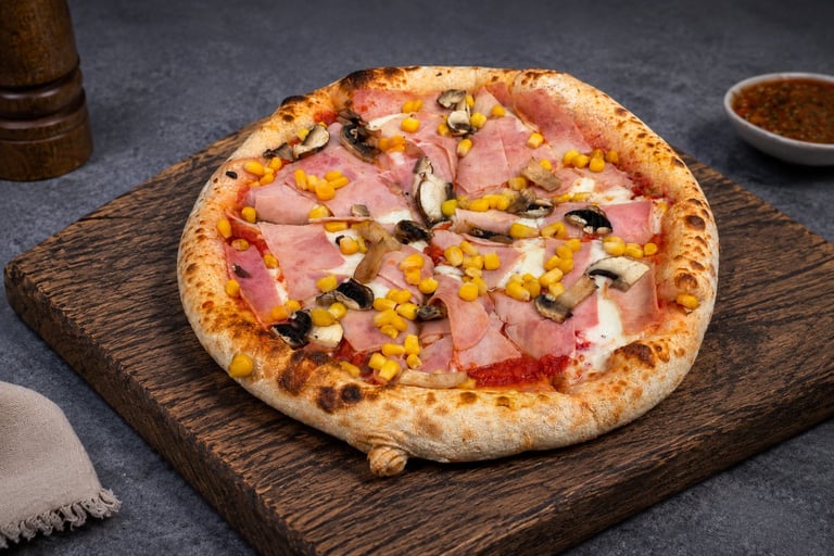 Wood-fired pizza topped with ham, corn, and black olives on a dark wooden board, served on a gray surface.