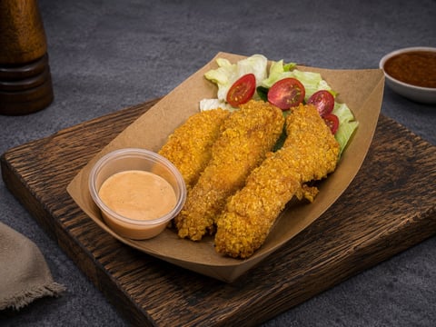 Golden fried chicken tenders on parchment paper with lettuce, tomato slices, and a side of dipping sauce on wooden board