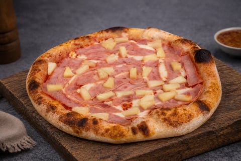 Freshly baked pizza with ham and pineapple on a wooden board, featuring a charred crust and melted cheese