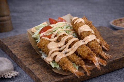 Fried shrimp skewers arranged on a wooden board with coleslaw, fresh tomato slices, and creamy sauce drizzle