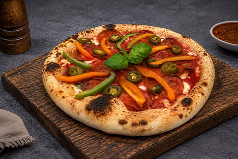 Artisan pizza with roasted red peppers, jalapenos, and basil on a wooden board, served on a dark surface