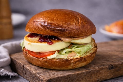 A gourmet sandwich on a toasted bun with melted cheese, tomato, lettuce, and red jam, served on a wooden board